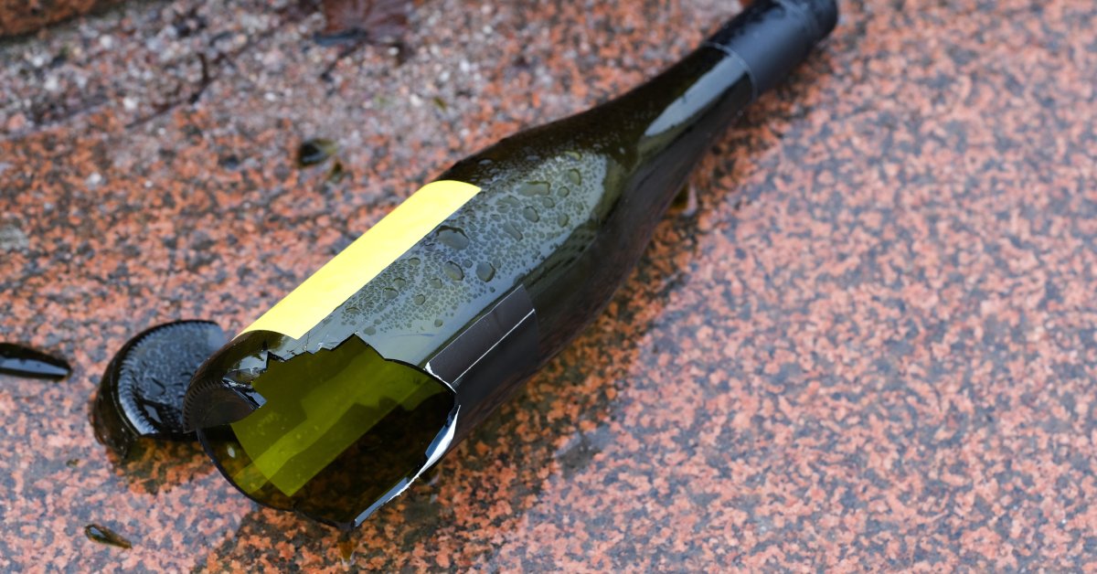 A close-up view shows a broken wine bottle lying on a red and black speckled surface. Wine surrounds the bottle.
