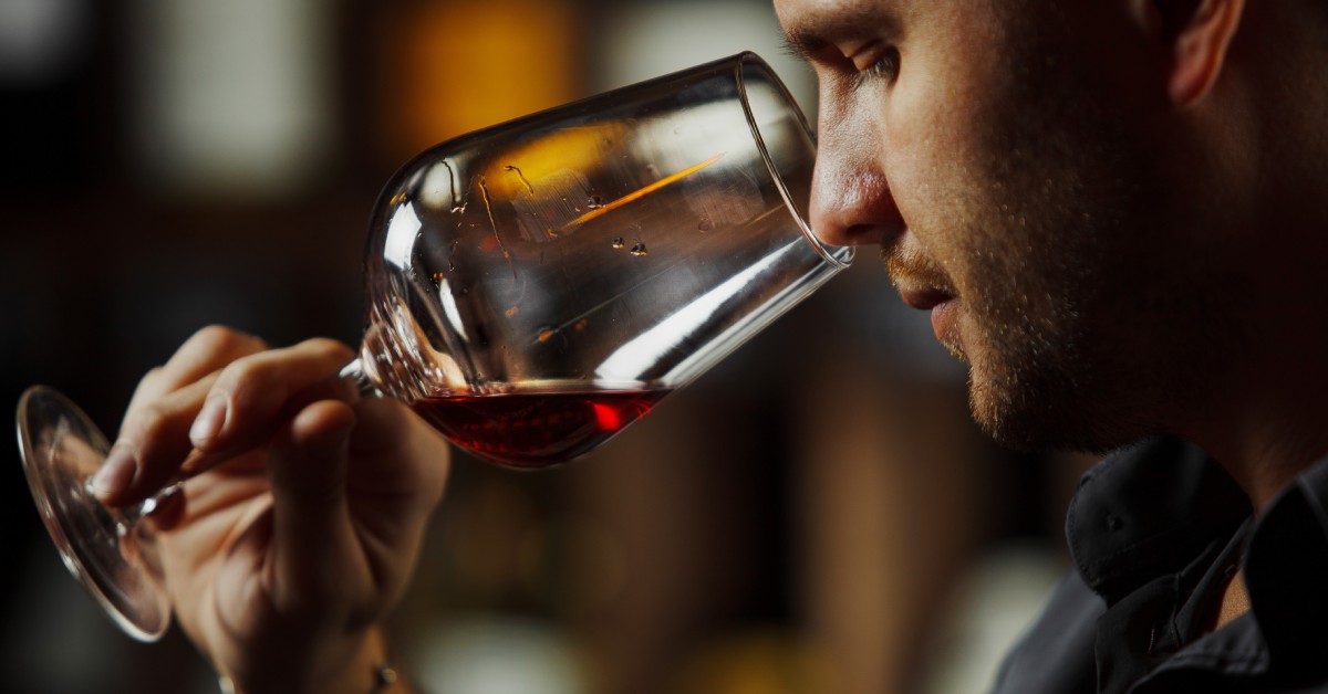 A close-up view shows a man holding a clear wine glass up to his nose. The glass contains red wine.