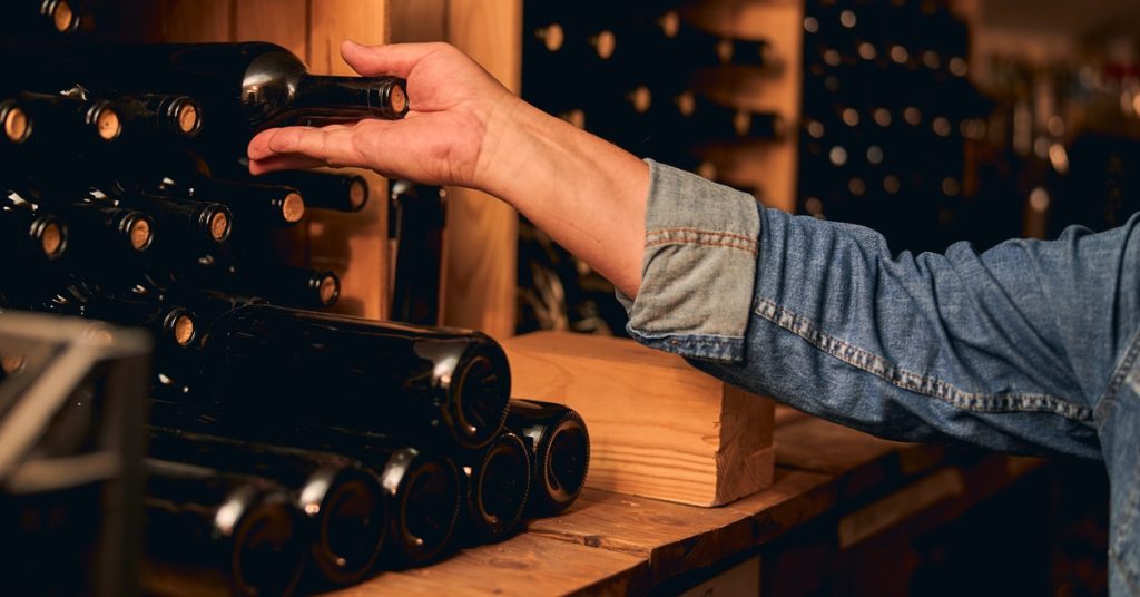 A person wearing a blue denim shirt with the sleeve rolled up reaches for a bottle of wine in a wine cellar.