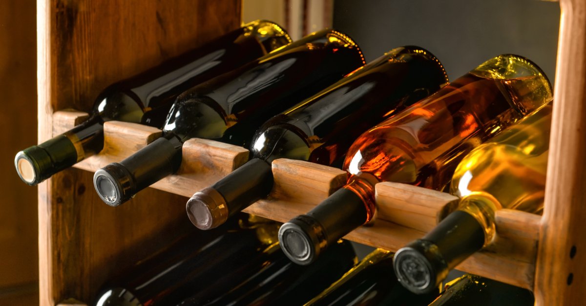 Ten bottles of wine without labels lay horizontally in a wine storage rack in a dimly-lit wine cellar.
