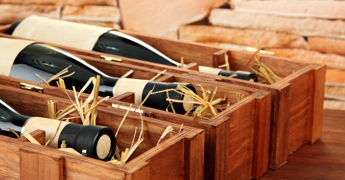 Three wine bottles with beige labels lie horizontally in individual wooden boxes filled with straw.