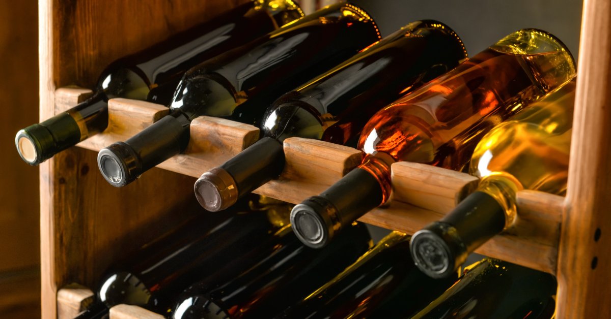 Ten unlabeled wine bottles lay horizontally on a wooden, two-level wine rack indoors.