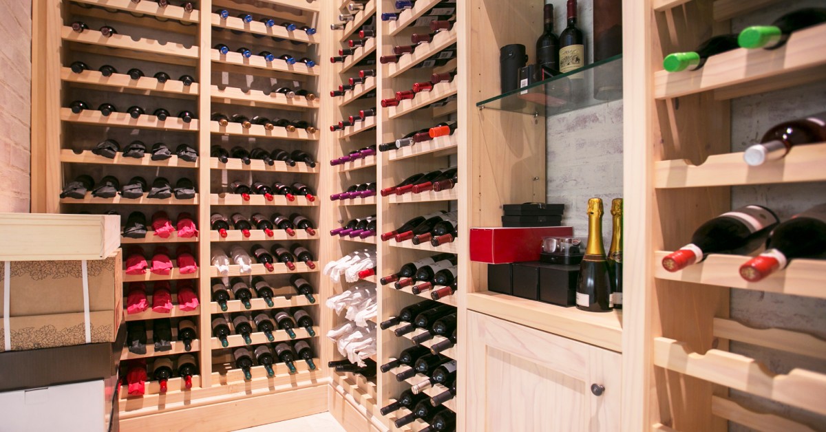 A brightly lit wine cellar with light, wooden shelving units. The shelves hold many horizontal wine bottles.
