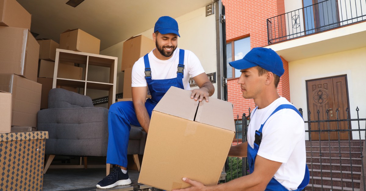 Two men in blue overalls and baseball caps lift a cardboard box into a moving truck full of boxes and furniture.