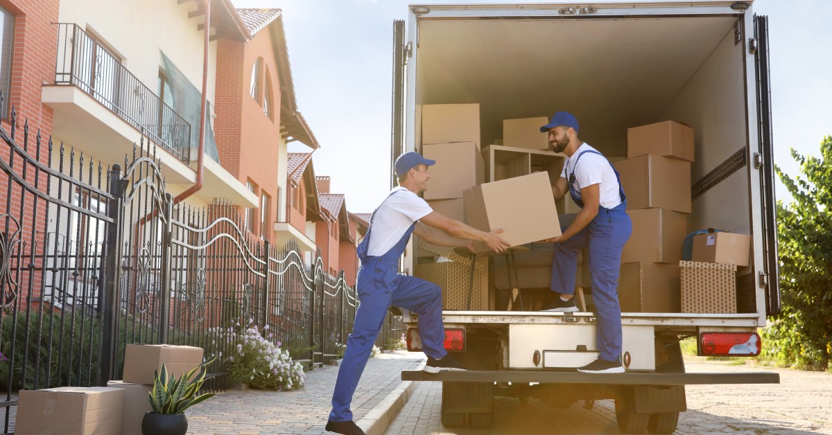 Two men in blue overalls and baseball caps work together to lift a cardboard box into a moving truck.