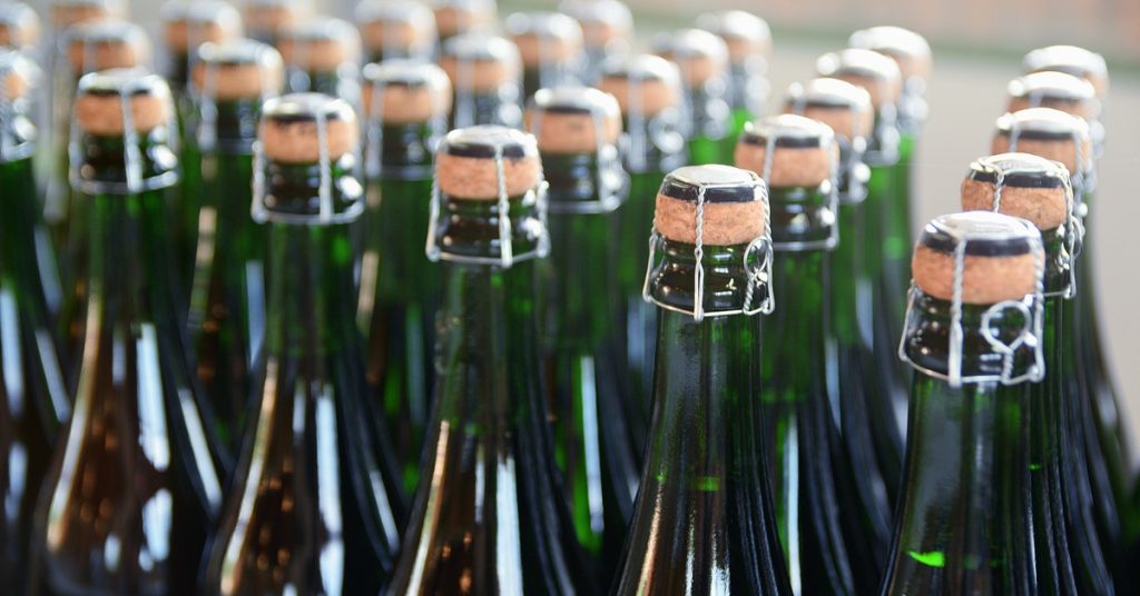 Many green champagne bottles with corks and metal tops are clustered and standing upright in a bright room.