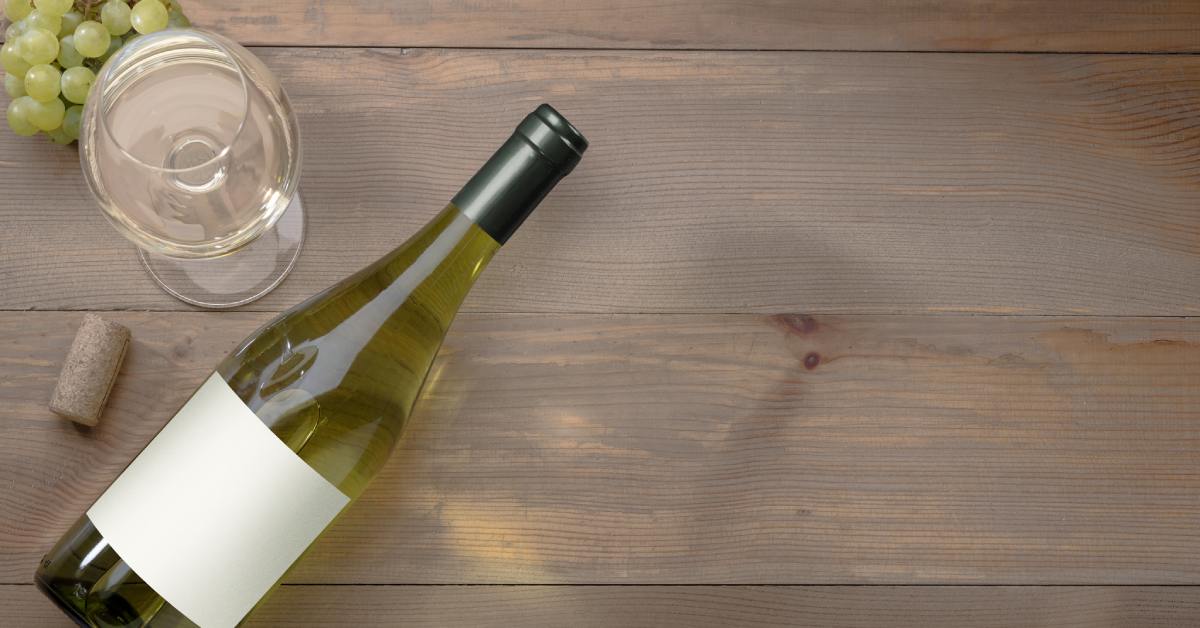 A bottle of chardonnay with a blank, white label on the bottle lying on a table next to a glass of white wine.