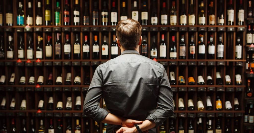 A man standing and surveying a large wine collection, with dozens of bottles arranged on dark brown wooden shelves.