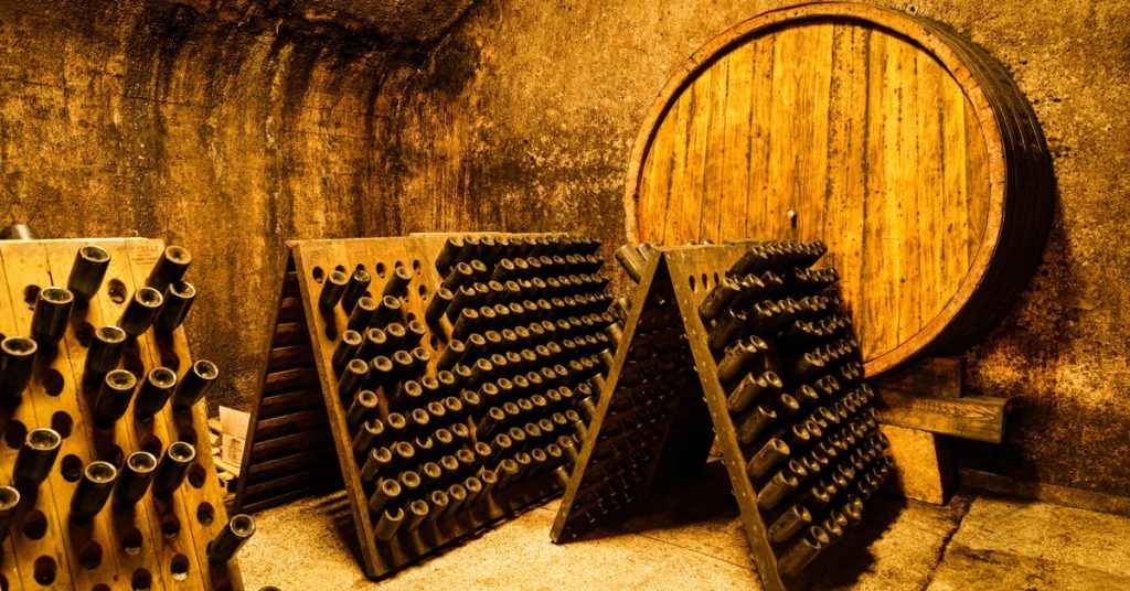 An old sparkling wine cellar that is warmly lit, with bottles held suspended in wine racks and a barrel in the background.