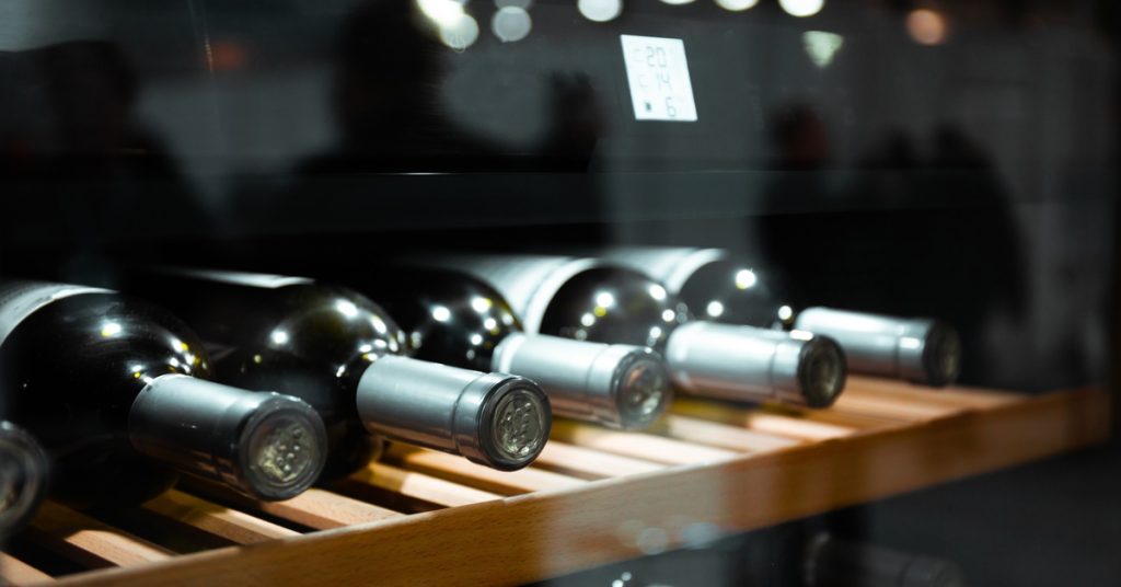 Bottles of wine on a wooden shelf under bright LED lighting, with another shelf full of wine just beneath it.